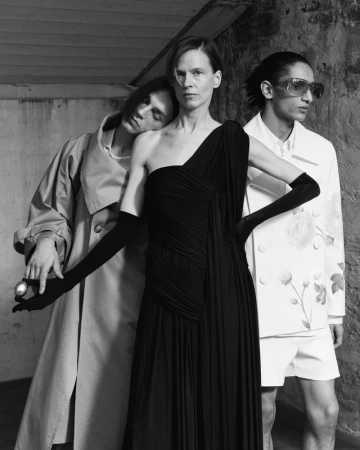 Three models posing in black and white fashion editorial, one wearing a pleated black gown and gloves while holding a metallic egg.