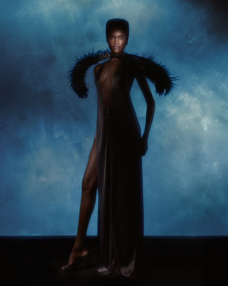 A fashionable adult female model poses indoors in a black gown with feather detail against a blue background in a studio fashion shot.