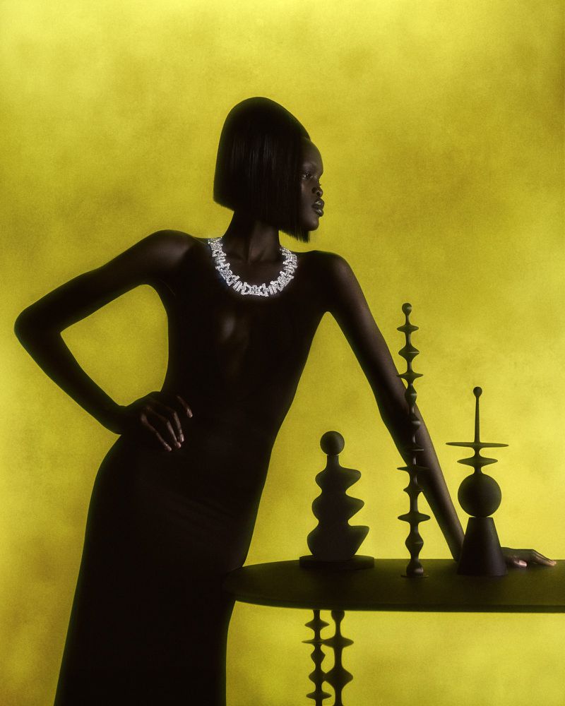 Elegant woman in a black dress and diamond necklace leans on a table with sculptural objects against a yellow textured backdrop.