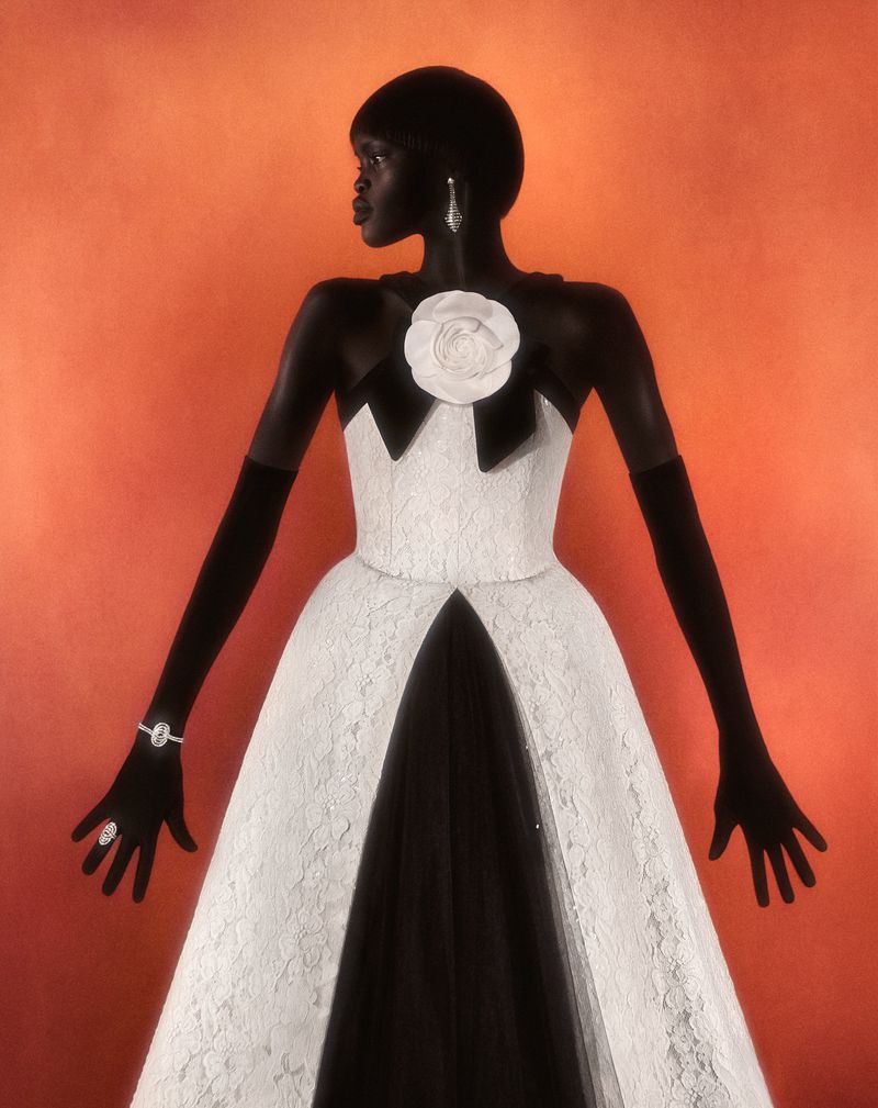 Elegant dark-skinned woman in a white lace gown with black accents posing in front of an orange backdrop, wearing jewelry and long gloves.