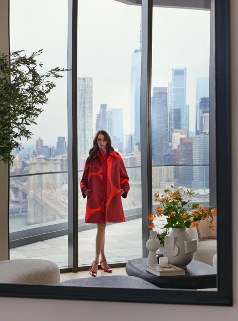 Fashionable woman in a patterned coat stands on a high-rise balcony with a stunning cityscape view, captured in a stylish, full-length shot.
