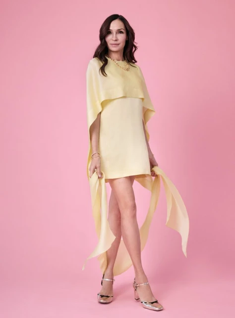 Elegant woman in yellow dress poses on pink backdrop. Fashionable female adult with long hair and stylish gold heels.