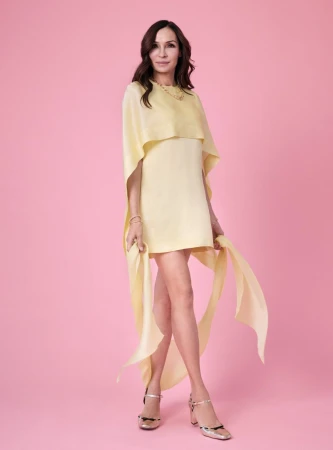 Elegant woman in yellow dress poses on pink backdrop. Fashionable female adult with long hair and stylish gold heels.
