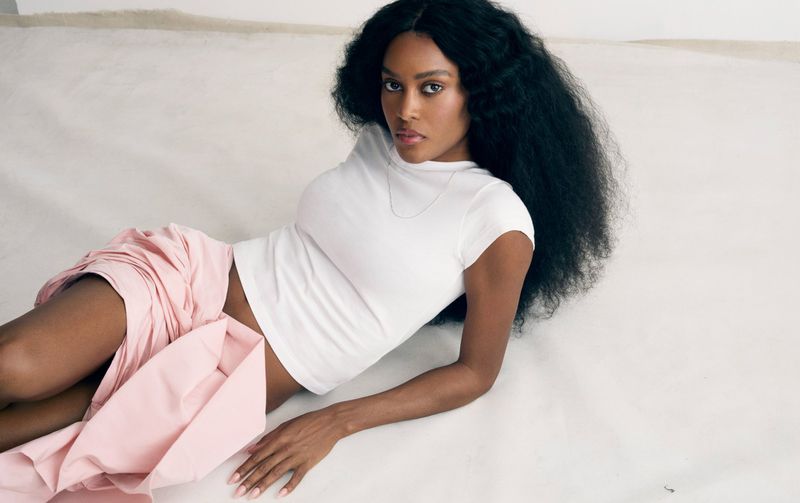 An adult woman poses while lying on a white surface wearing a white t-shirt and pink skirt, showcasing fashion and beauty.