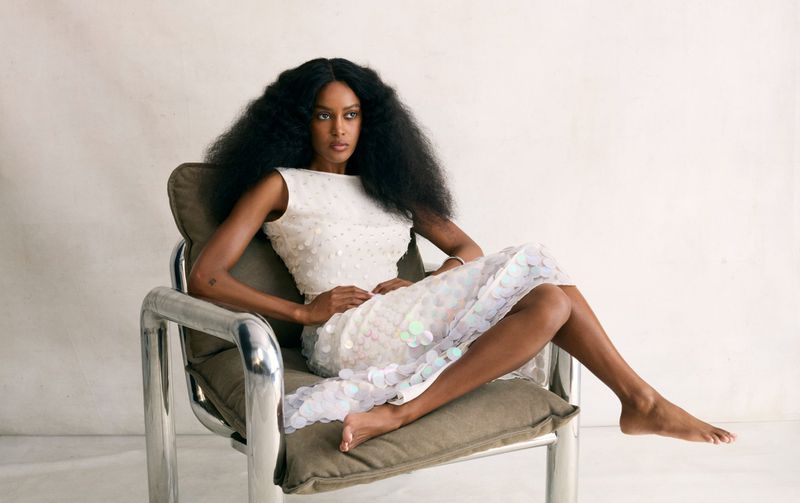 Elegant African-American woman in a white dress with sequins, barefoot, sitting in a metal-framed chair in a light studio setting.