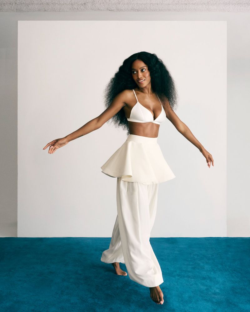 A barefoot Black woman in a white bralette and wide-leg pants dances on a teal floor against a white backdrop in a studio setting.