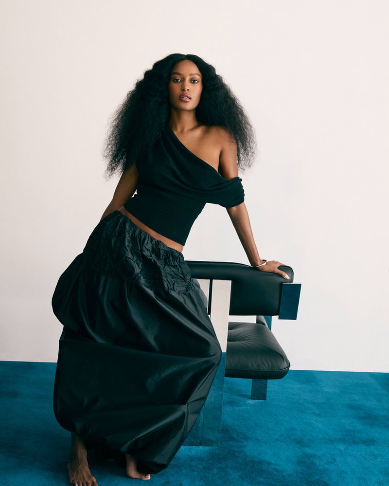Adult woman with dark curly hair poses in a black top and large black skirt, leaning on a chair in a brightly lit indoor setting.