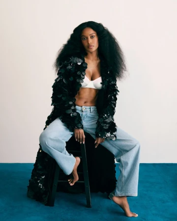 A barefoot Black woman with dark curly hair sits on a stool wearing blue jeans, a bra, and a sequined black coat in a studio setting.