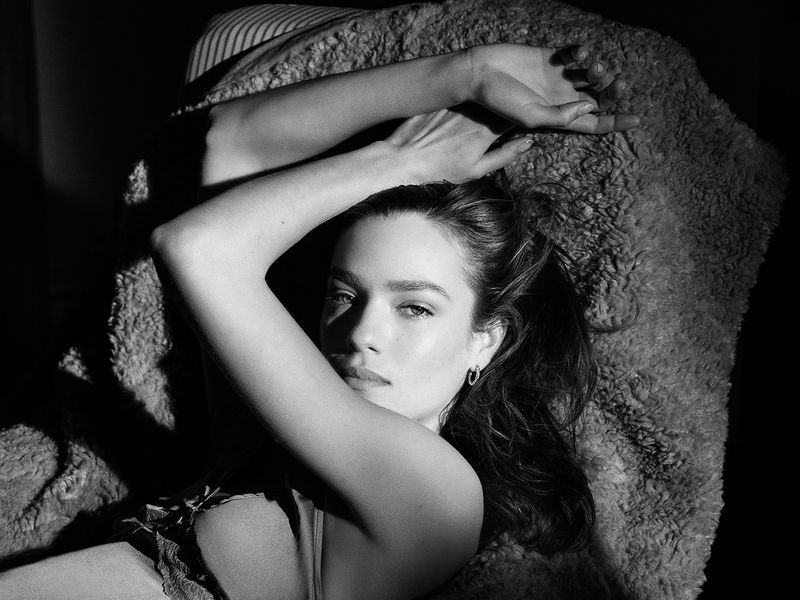 Monochrome portrait of a woman with arms raised overhead, reclining on a textured blanket in a dimly lit indoor setting.