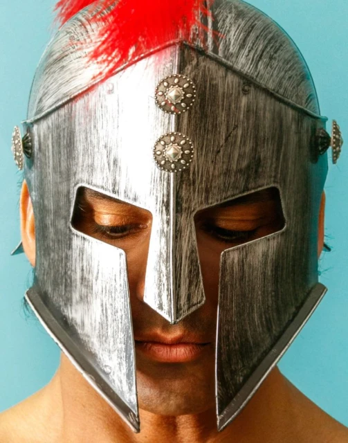 Close-up of an adult male wearing a silver Roman helmet with red plumes against a light blue background, looking down with neutral expression.
