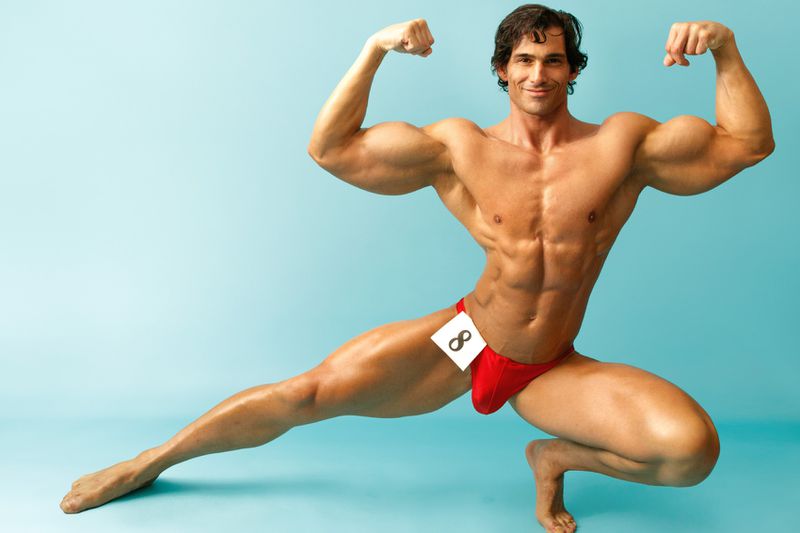 Muscular man flexing in a red posing suit, studio shot against a blue background, showing off physique and athletic conditioning.
