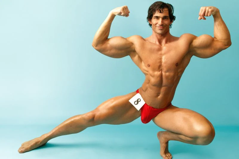 Muscular man flexing in a red posing suit, studio shot against a blue background, showing off physique and athletic conditioning.