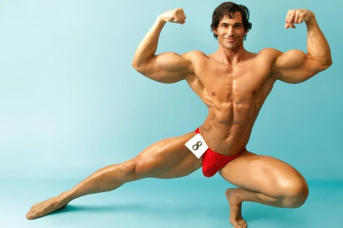 Muscular man flexing in a red posing suit, studio shot against a blue background, showing off physique and athletic conditioning.