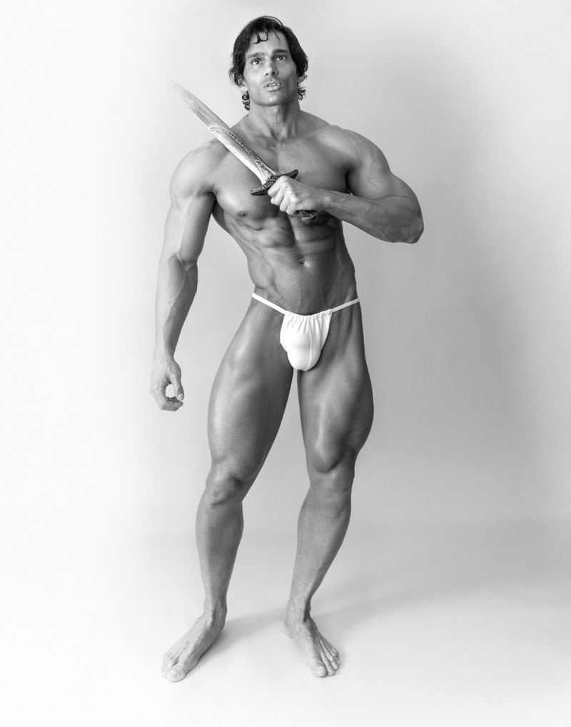 Black and white portrait of muscular man holding a knife. The man is shirtless with minimal clothing in a bright, neutral environment.