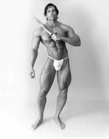 Black and white portrait of muscular man holding a knife. The man is shirtless with minimal clothing in a bright, neutral environment.