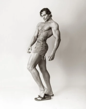Muscular man posing in profile wearing denim cut-off shorts and flip-flops against a white studio background.