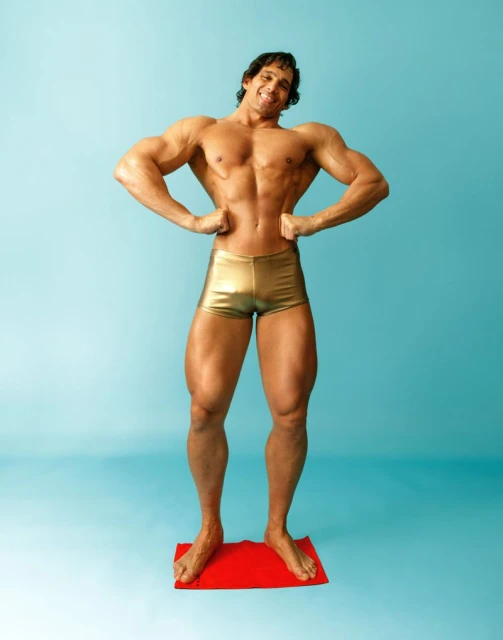 Muscular man with bronze skin and gold shorts stands on a red mat. He's posing against a blue background, showcasing defined abdominal muscles.