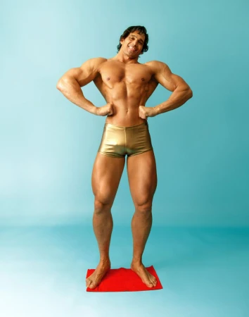 Muscular man with bronze skin and gold shorts stands on a red mat. He's posing against a blue background, showcasing defined abdominal muscles.