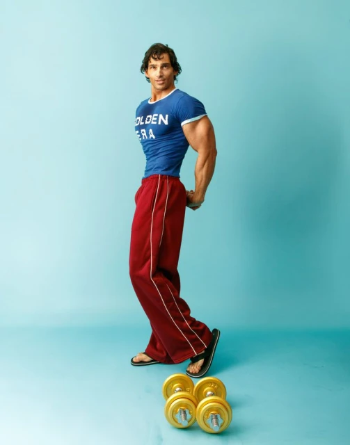 Muscular man in blue t-shirt and maroon pants posing in a photo studio next to gold-colored dumbbells on an aqua backdrop.