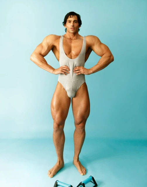 Full body studio portrait of a muscular man wearing a leotard, standing with hands on waist, and push-up bars at his feet, on blue backdrop.