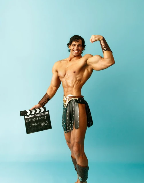 Muscular man in ancient-style costume strikes a pose holding a clapboard on a pale blue background, flexing his biceps.