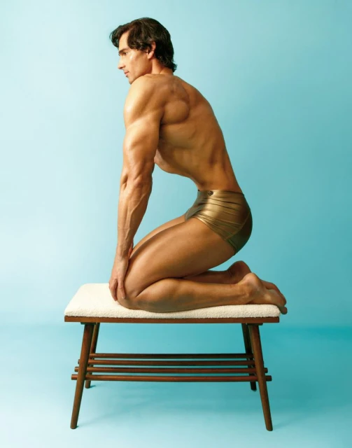 A muscular adult male kneeling on a stool in a studio setting, wearing gold shorts, photographed against a light blue backdrop.
