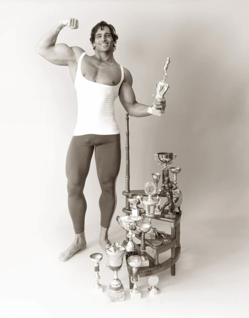 A physically fit adult male stands with a vintage trophy collection, flexing his bicep in an indoor photo studio against a neutral backdrop.