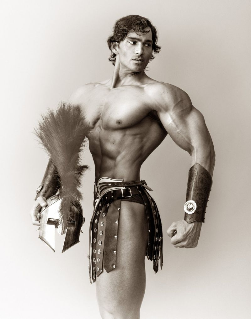 Muscular man in Roman-style armour poses indoors with a helmet and feathered plume; black and white portrait.