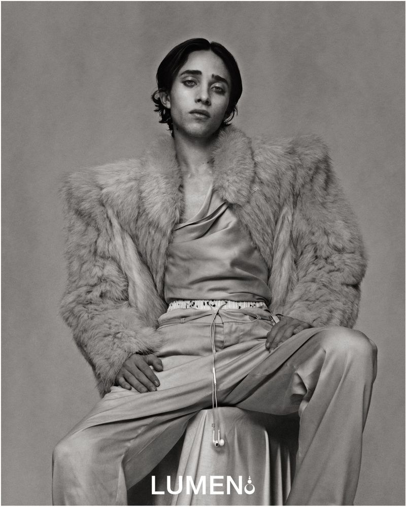 Monochrome portrait of a person with long dark hair, wearing a satin shirt and a fur coat, seated indoors against a grey background.