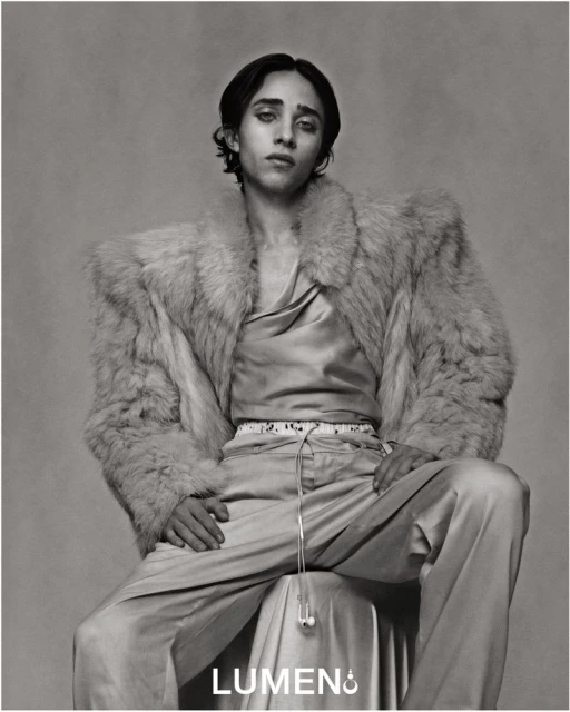 Monochrome portrait of a person with long dark hair, wearing a satin shirt and a fur coat, seated indoors against a grey background.