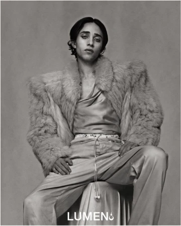 Monochrome portrait of a person with long dark hair, wearing a satin shirt and a fur coat, seated indoors against a grey background.