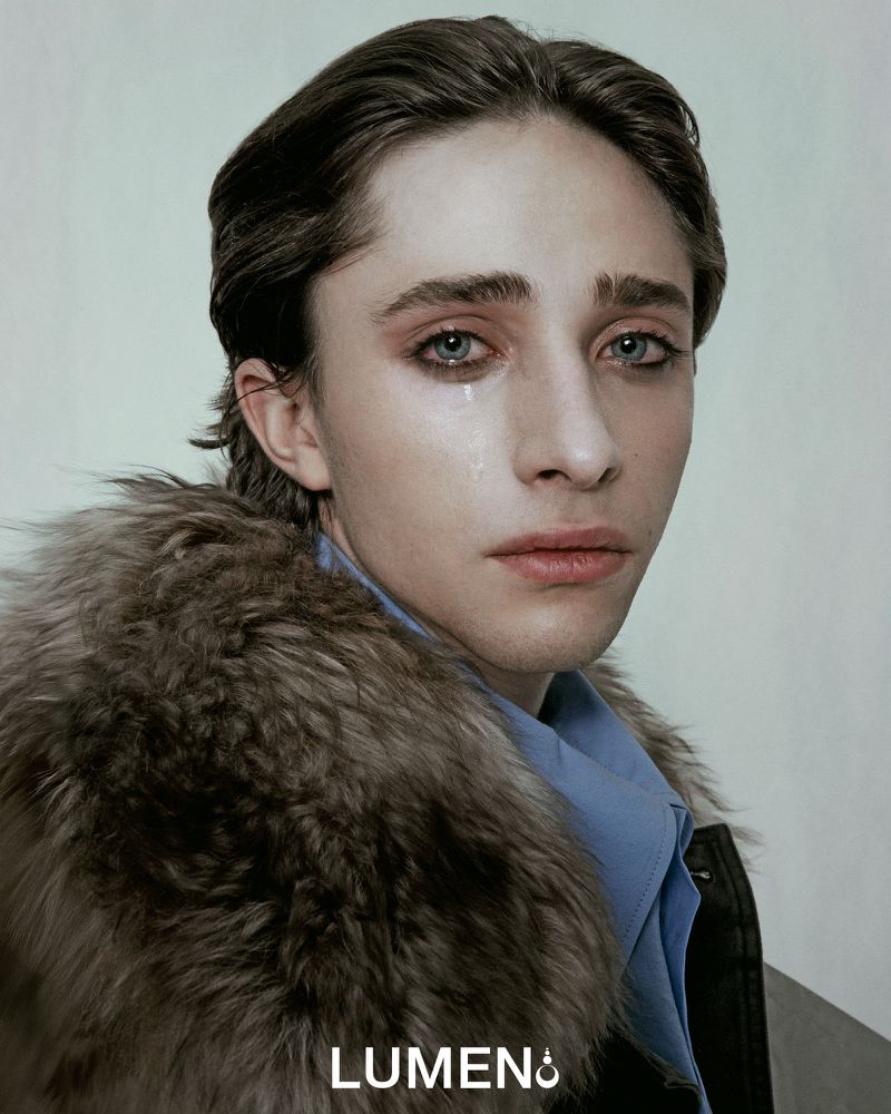 Close-up studio shot of a young adult with tear, fur-trimmed jacket, and light blue shirt against a neutral background, showcasing fashion and beauty.