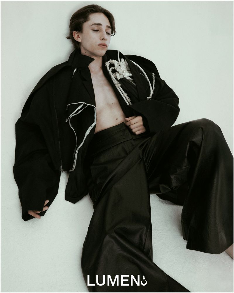 Young adult male fashion model in black jacket and wide-leg leather pants poses against a light wall in a studio setting.