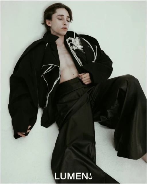 Young adult male fashion model in black jacket and wide-leg leather pants poses against a light wall in a studio setting.