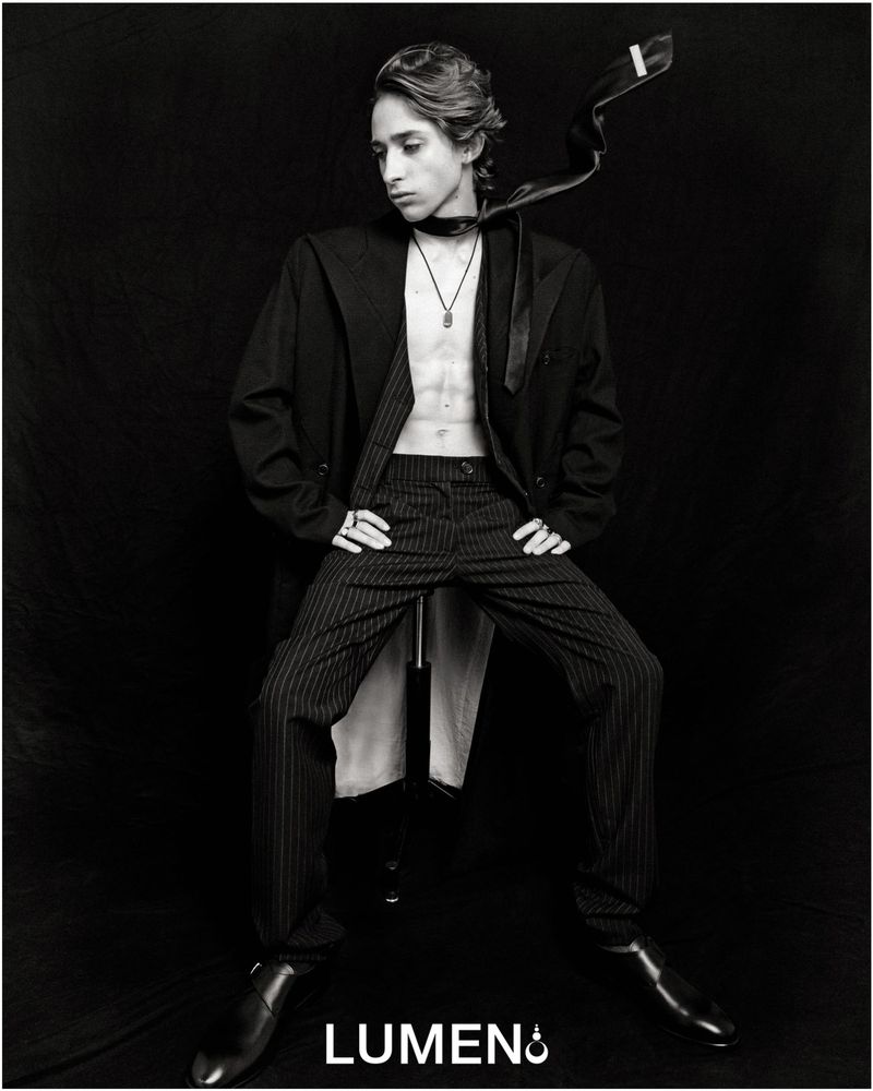 Monochrome portrait of a young adult with an unbuttoned jacket, striped pants, and shiny shoes against a dark background.