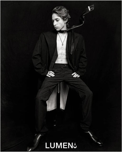 Monochrome portrait of a young adult with an unbuttoned jacket, striped pants, and shiny shoes against a dark background.