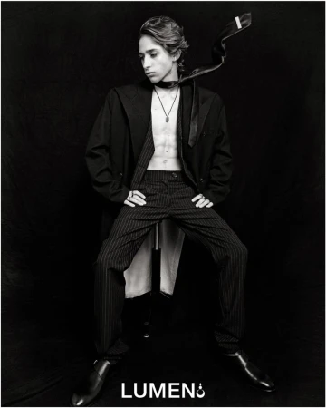 Monochrome portrait of a young adult with an unbuttoned jacket, striped pants, and shiny shoes against a dark background.