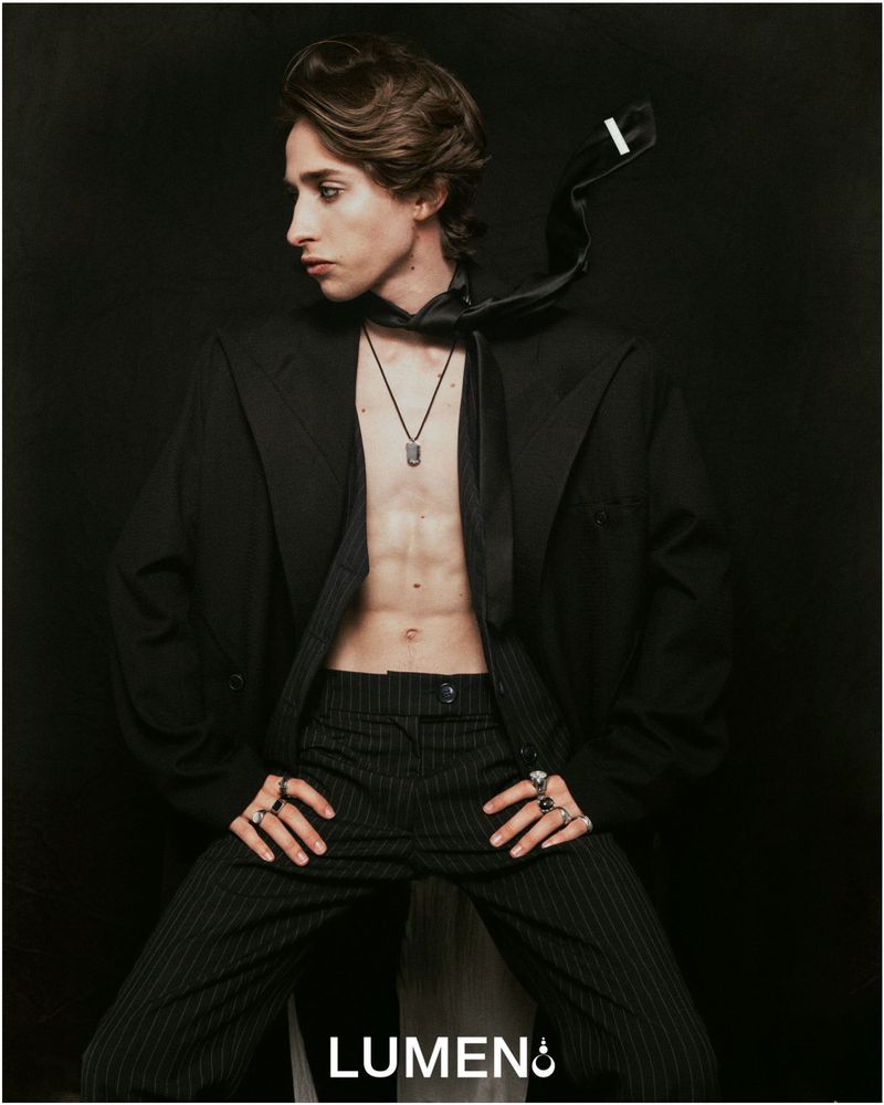 A young man with dark eyeliner wears an unbuttoned black pinstripe suit jacket and matching trousers, posing against a solid black background.