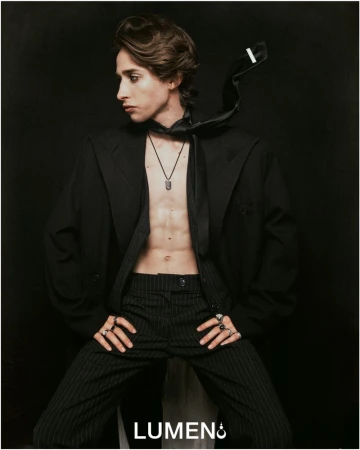 A young man with dark eyeliner wears an unbuttoned black pinstripe suit jacket and matching trousers, posing against a solid black background.