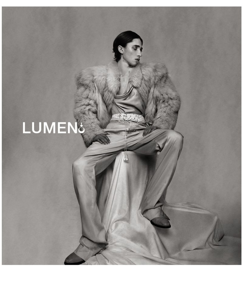 Monochrome portrait of a stylish adult male in a fur coat, satin pants, and shoes, seated on a draped fabric surface against a gray backdrop.