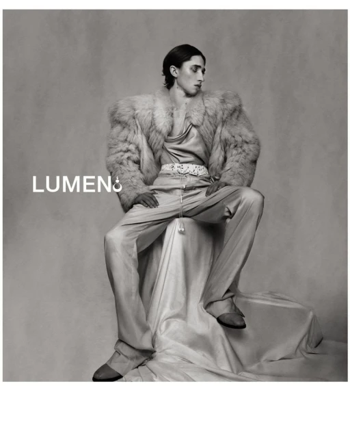 Monochrome portrait of a stylish adult male in a fur coat, satin pants, and shoes, seated on a draped fabric surface against a gray backdrop.