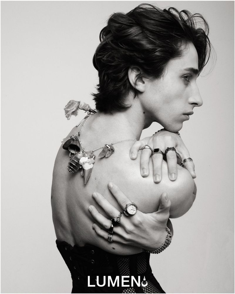 Monochrome portrait of a shirtless person with dark hair, adorned with rings and a floral necklace, posing against a stark white backdrop.