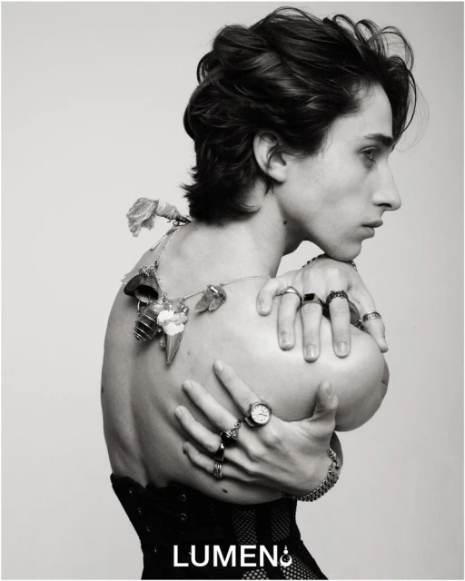 Monochrome portrait of a shirtless person with dark hair, adorned with rings and a floral necklace, posing against a stark white backdrop.