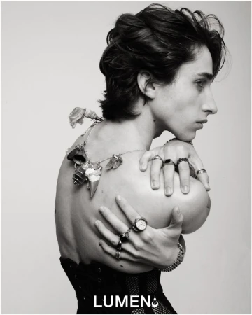 Monochrome portrait of a shirtless person with dark hair, adorned with rings and a floral necklace, posing against a stark white backdrop.