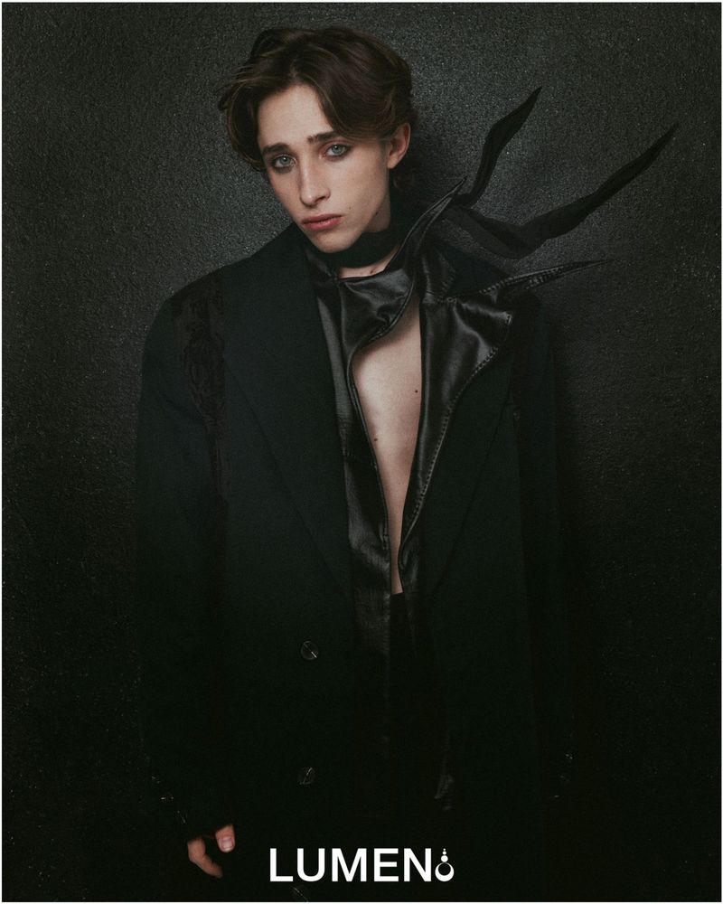Portrait of a young adult male in a black coat and neckpiece against a dark background, showcasing fashion and studio lighting.