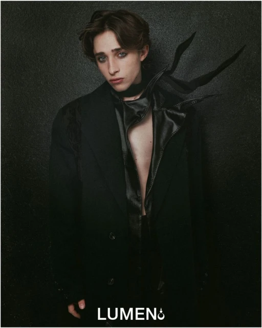 Portrait of a young adult male in a black coat and neckpiece against a dark background, showcasing fashion and studio lighting.