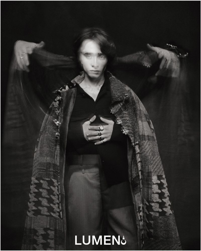 Monochrome portrait of a woman with long exposure effect, wearing a patterned coat and rings, against a dark background in a studio.