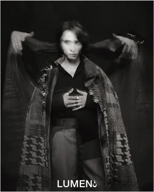 Monochrome portrait of a woman with long exposure effect, wearing a patterned coat and rings, against a dark background in a studio.