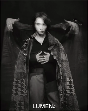 Monochrome portrait of a woman with long exposure effect, wearing a patterned coat and rings, against a dark background in a studio.