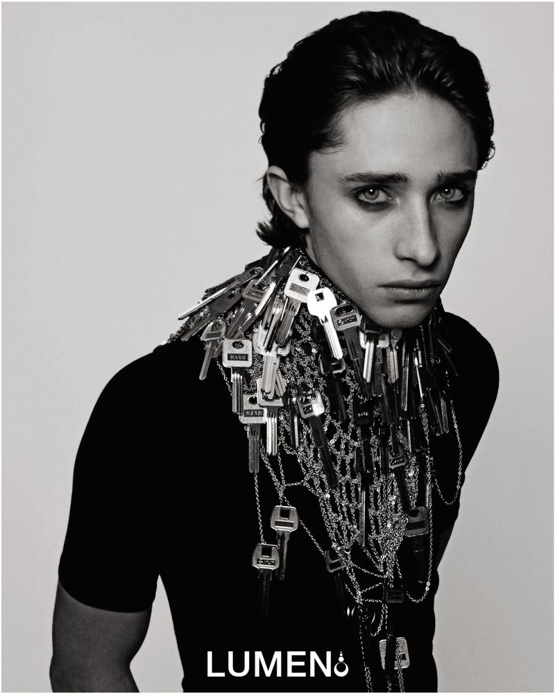 Monochrome portrait of a young adult wearing a black top and an elaborate key necklace against a neutral backdrop.